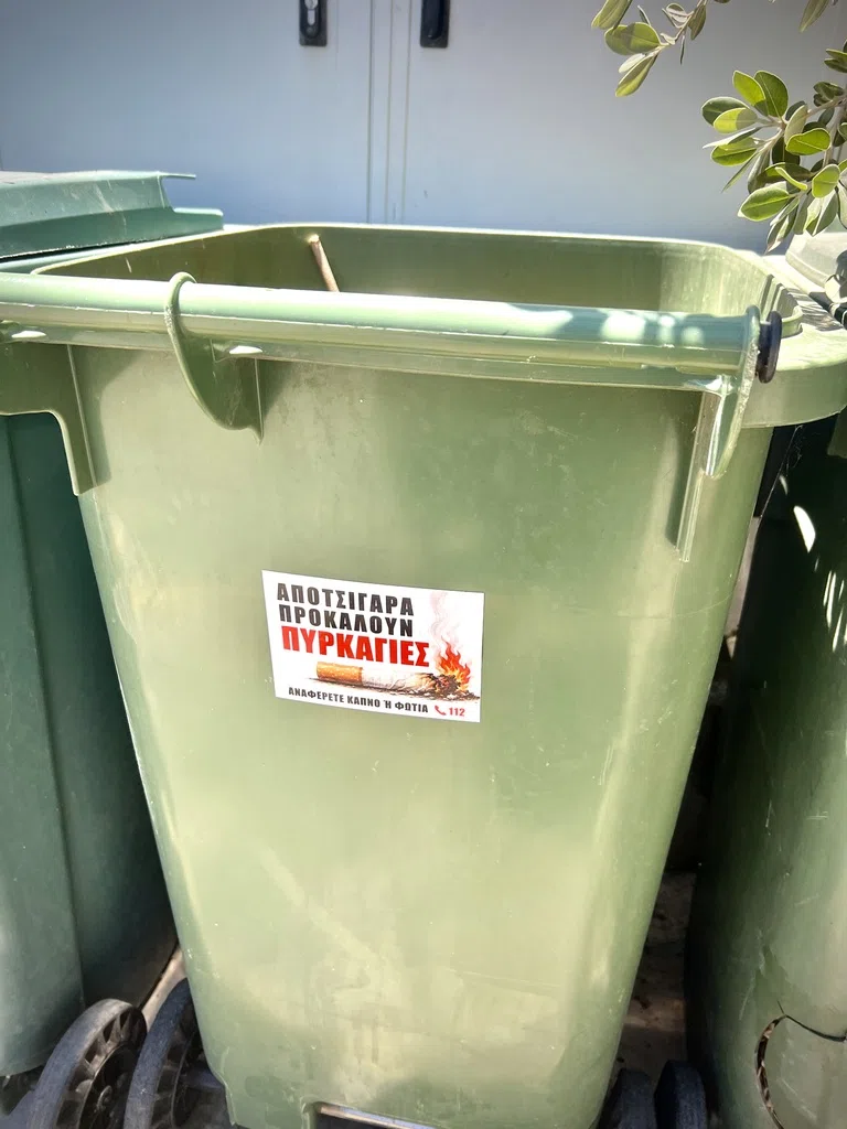 A Greek cigarette-butt fire warning sticker on a green bin